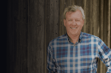 1000 Stories Winemaker, Bob Blue, stands in front of a wooden backdrop wearing a blue plaid button up shirt.